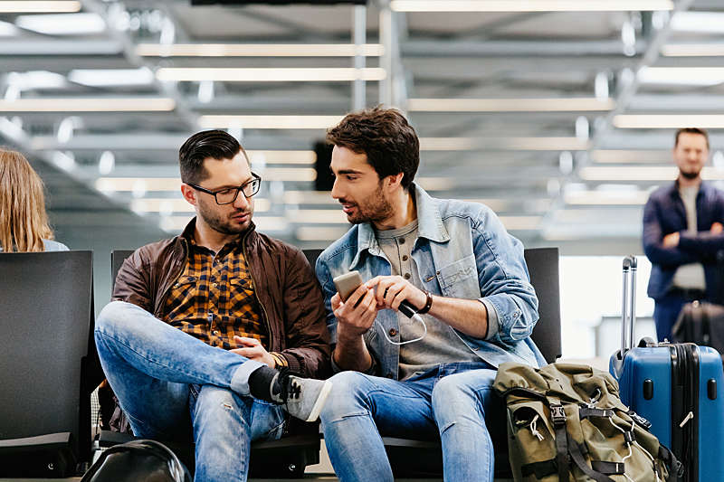 Two young men sitting at an airport lounge using a mobile phone.