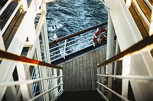 Flight of stairs descending towards guard rail of cruise ship with gull perched on top and ocean visible in background.