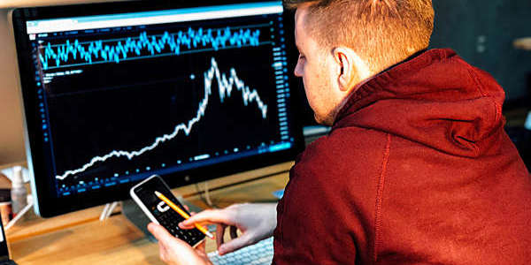 Man follows a stock chart on a computer screen while tapping away on his mobile phone.