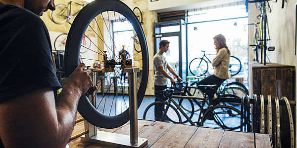 Close shot of bicycle mechanic repairing a wheel while a salesman attends a client in the background.