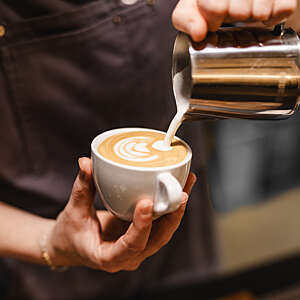 Close up view of the hands of a barista preparing a coffee by drawing a flower with milk.